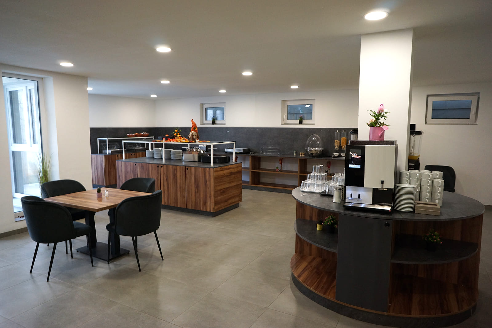 Speisesaal mit Buffetinsel, Kaffeevollautomat und Sitzplätzen an Holztischen.