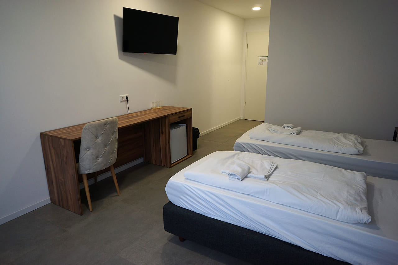 Langer Schreibtisch mit Stuhl und Minibar gegenüber dem Bett, darüber ein Wandfernseher.