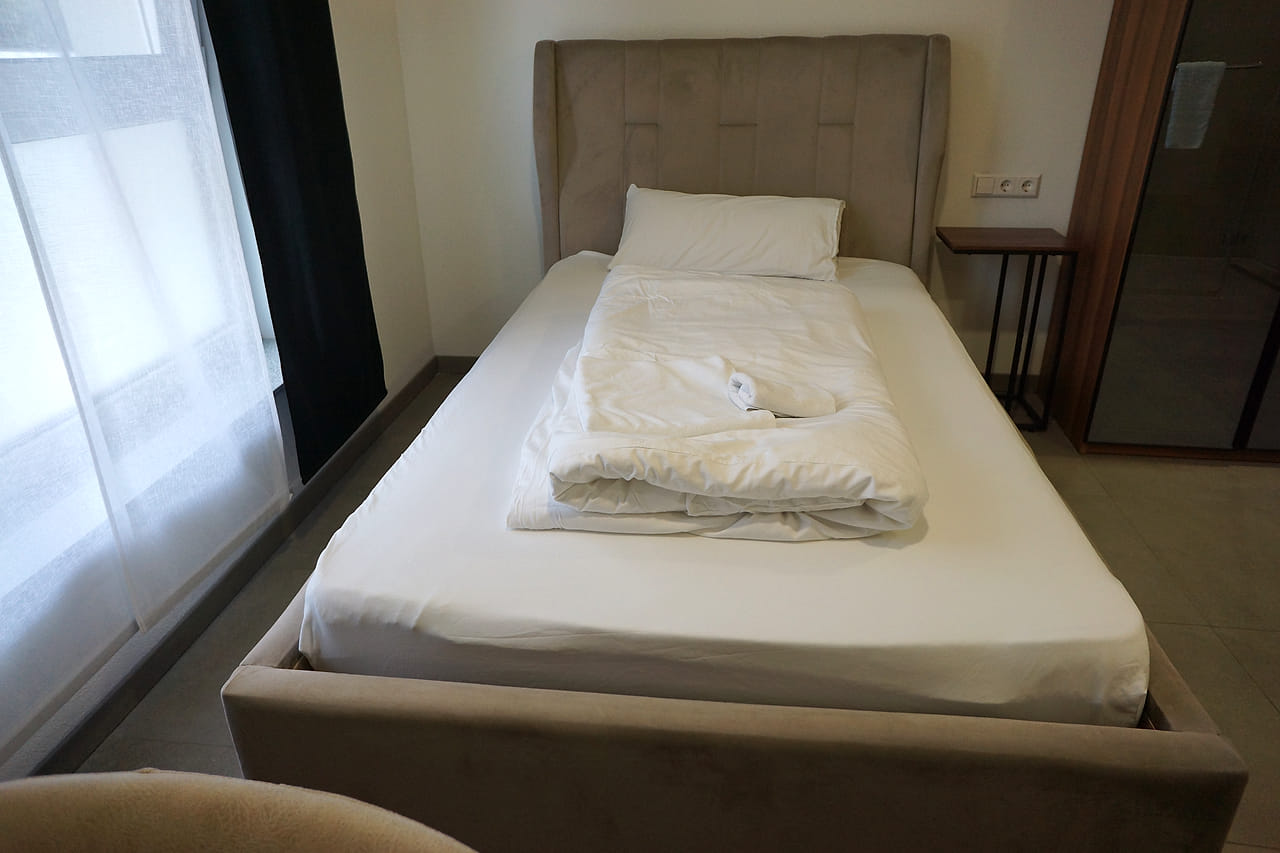 Einzelbett mit gepolstertem Kopfteil vor dem Fenster, daneben ein Nachttisch.