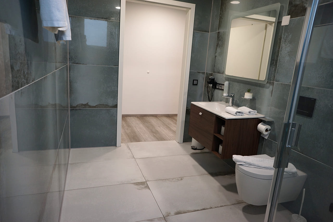 Großes Bad des Studio Apartments mit grün marmorierten Fliesen, Dusche hinter Glas und beleuchtetem Spiegel.